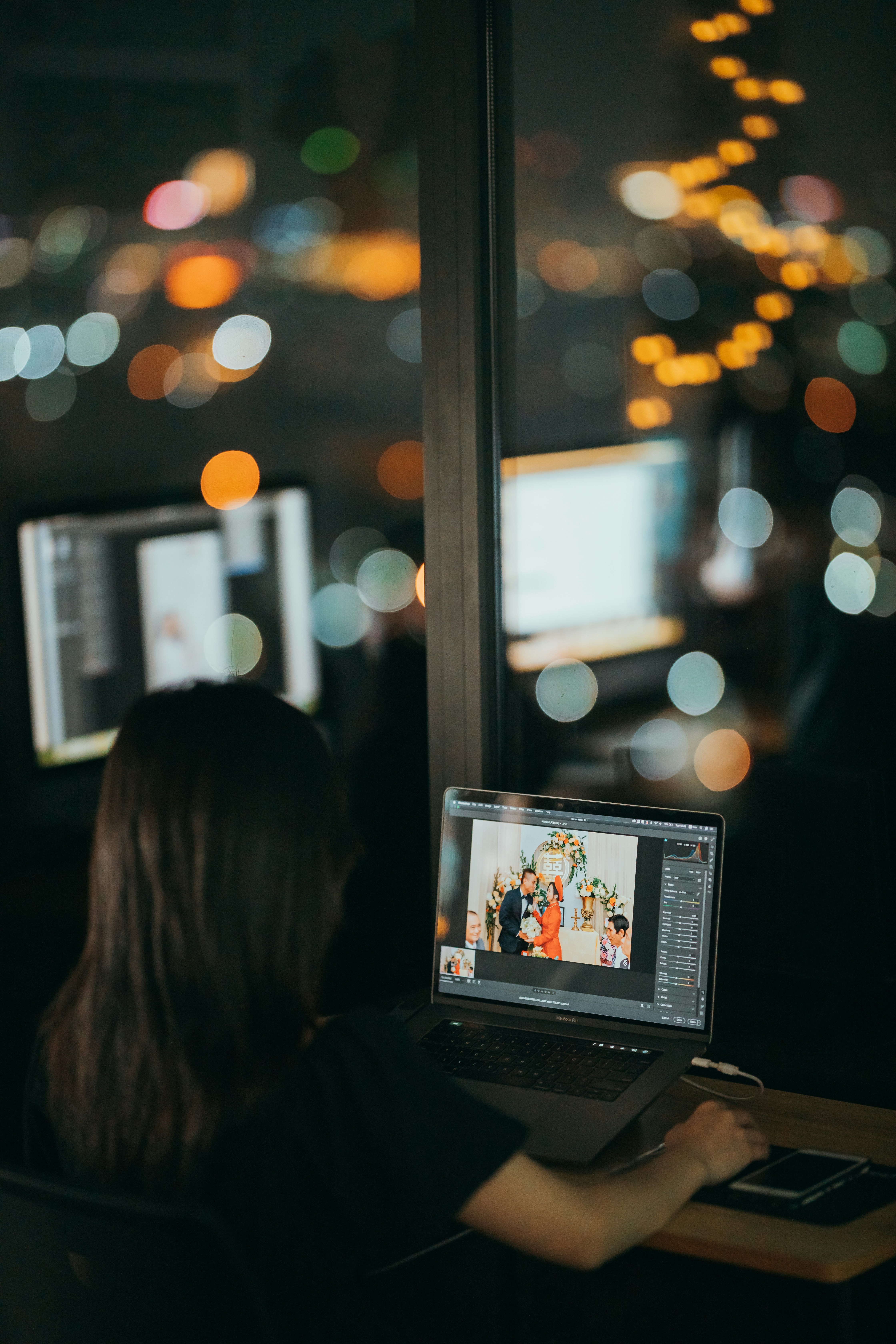 A woman with long dark hair sits with her back to the camera.  She is looking at a computer monitor where editing software is displayed