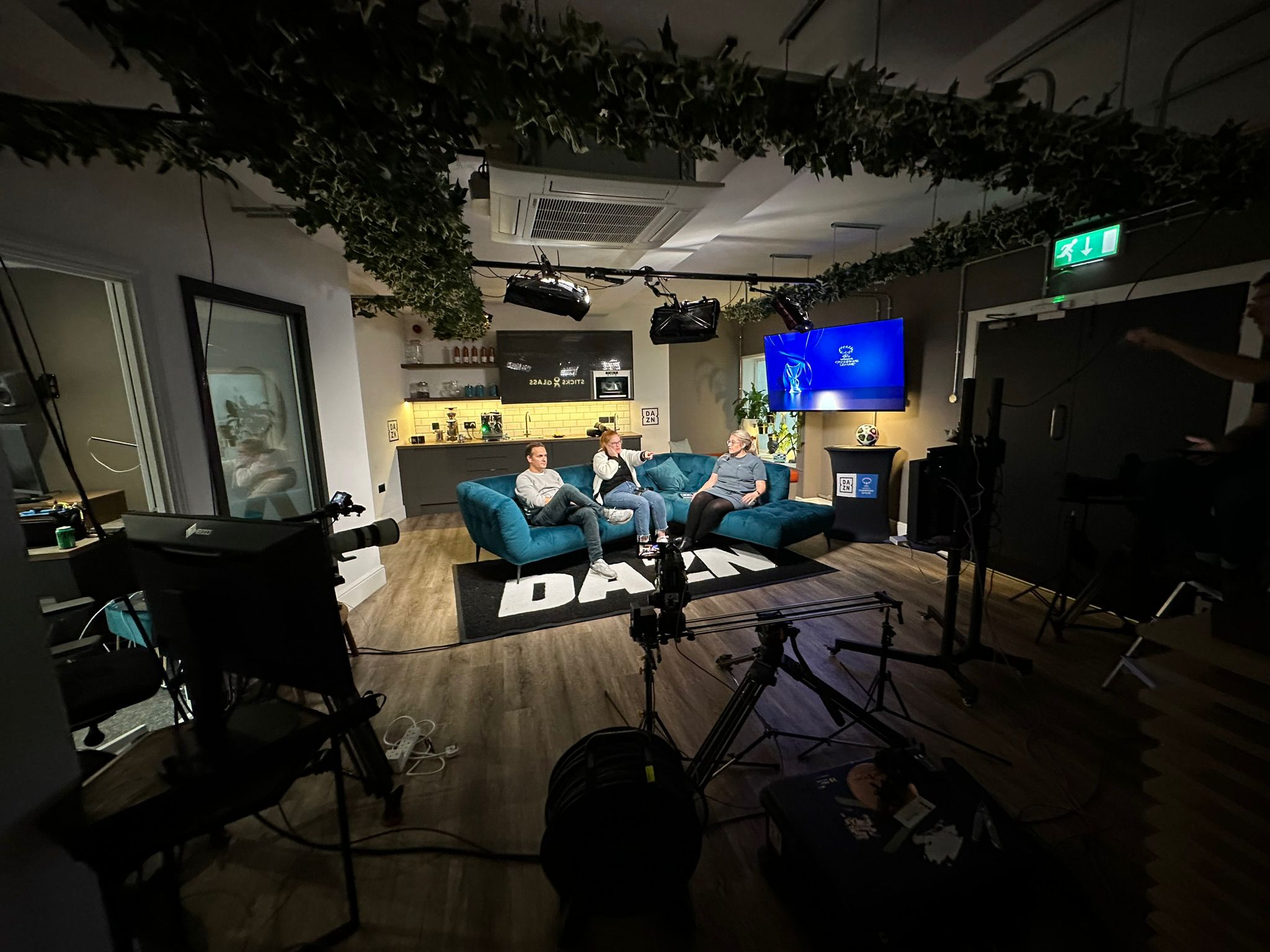 A podcast studio filled with equipment. In the centre three presenters sit together on a sofa