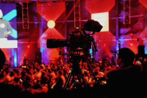 A camera operator working with a large camera. A seated studio audience can be seen in the background.