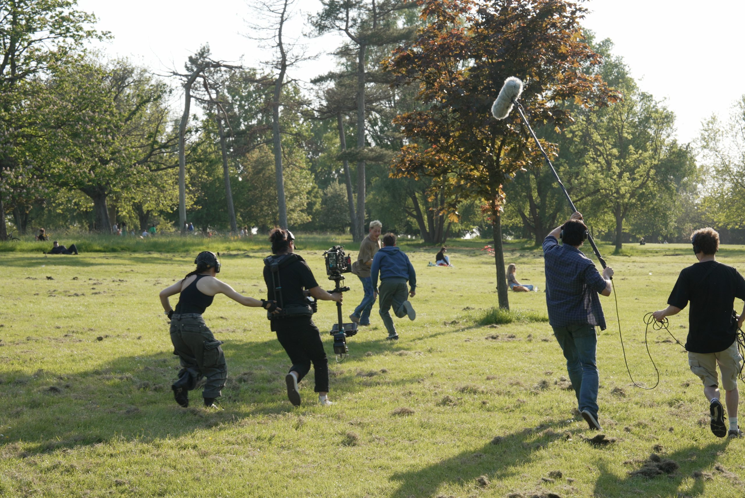 A film crew shoots an outdoor scene featuring two actors running through a park. The crew carry boom mics and a steadicam.