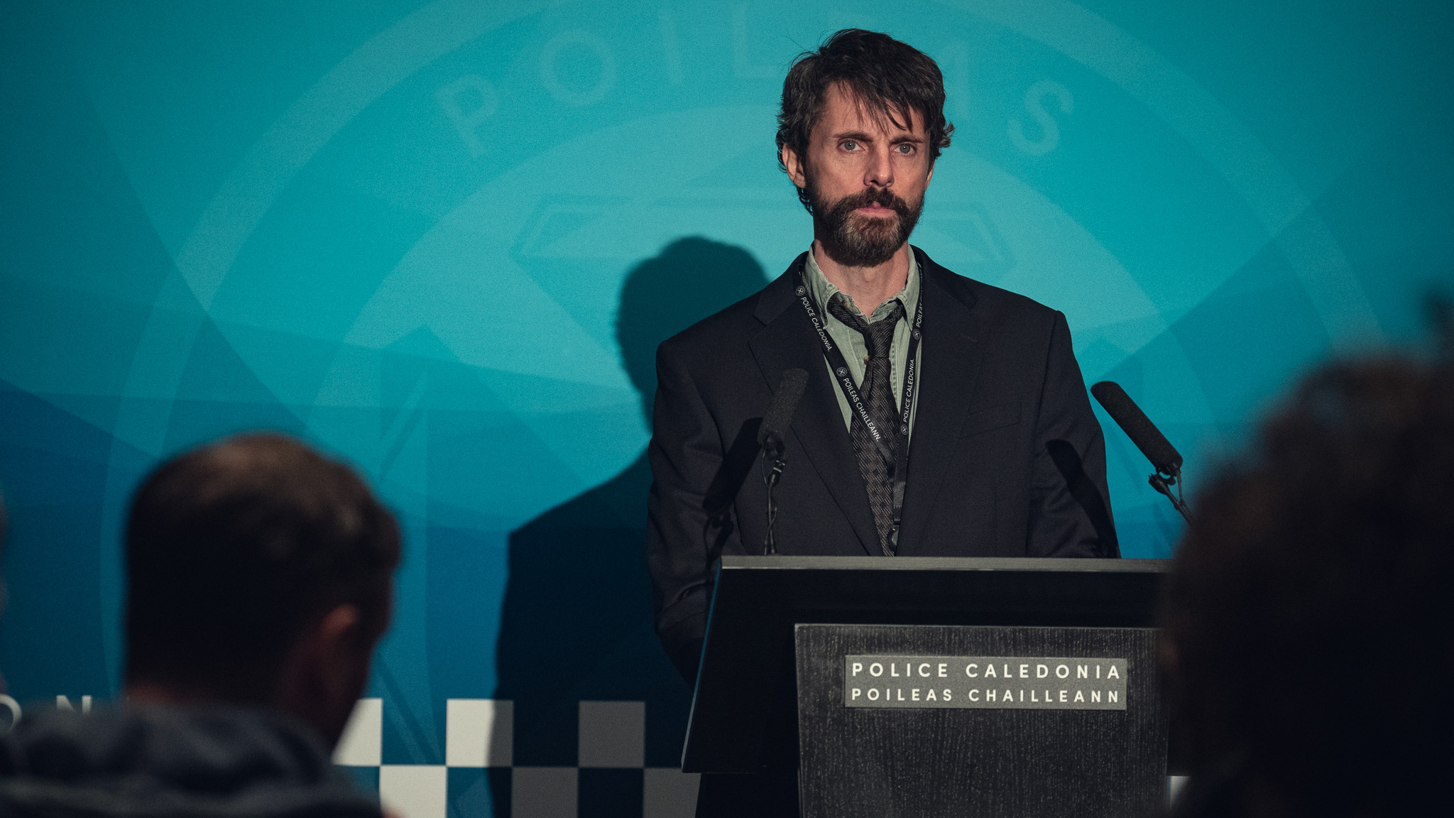 Still from Netflix series Dept Q featuring lead actor Matthew Goode as character Det Carl Morck appearing at a news conference, standing behind a lectern brabded Police Caledonia