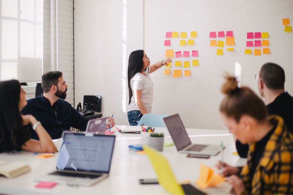 A team is having a meeting around a table. Each member has a laptop in front of them. The team leader points to a board filled with coloured Post-It notes.
