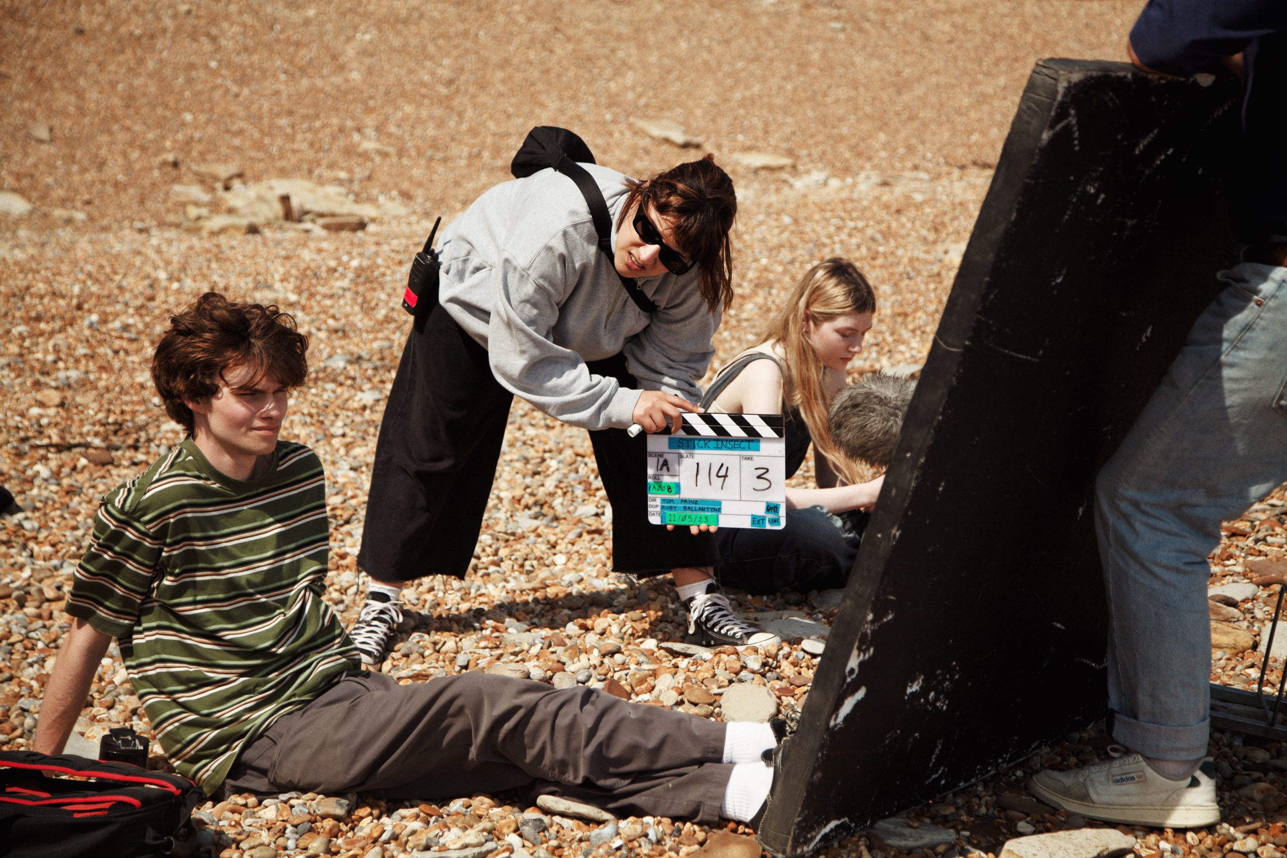 A pair of actors sat on a pebble beach with a member of the production crew hitting a clapperboard.