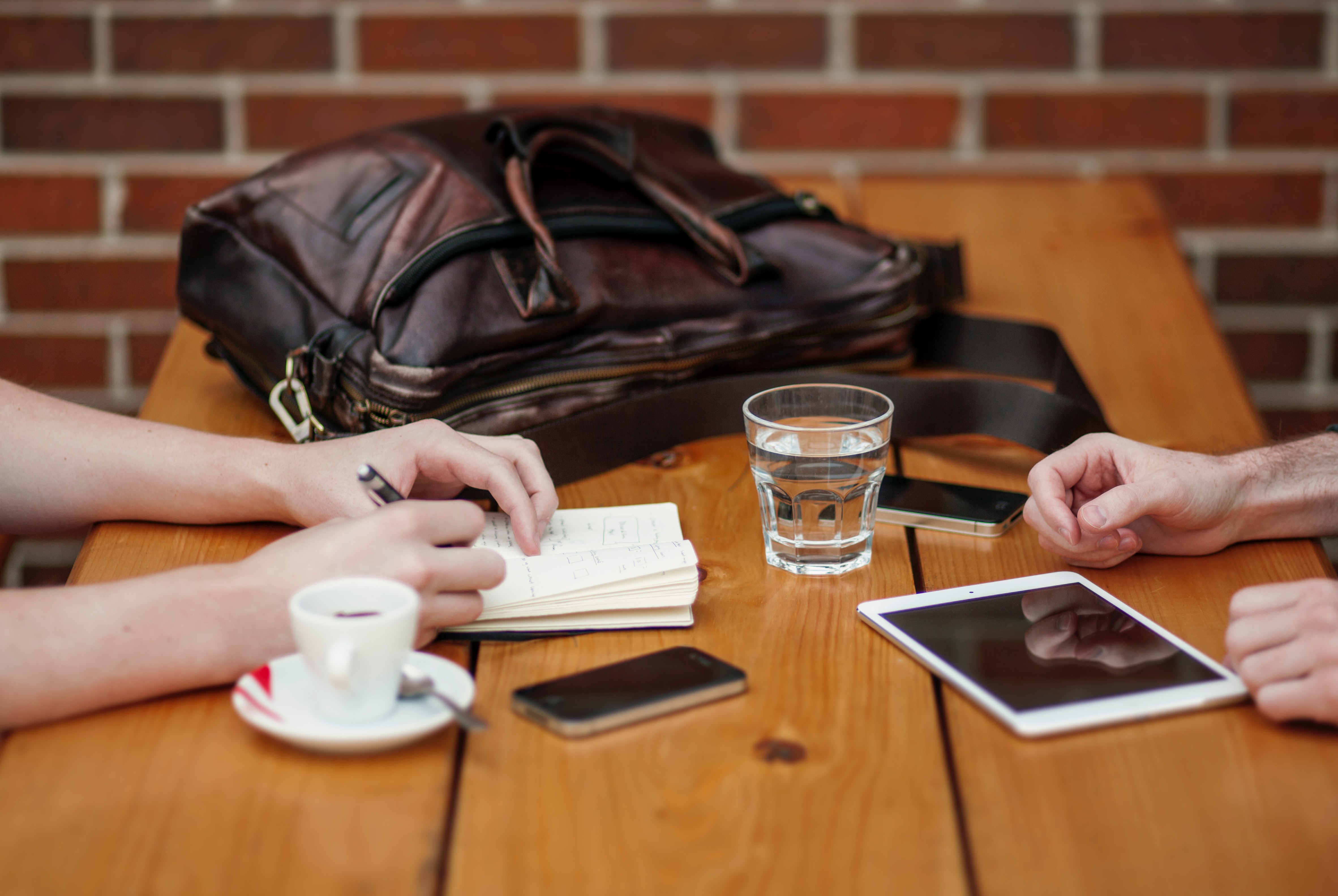 Two people sitting down with only their arms showing. Notepad, tablet and drinks on the table