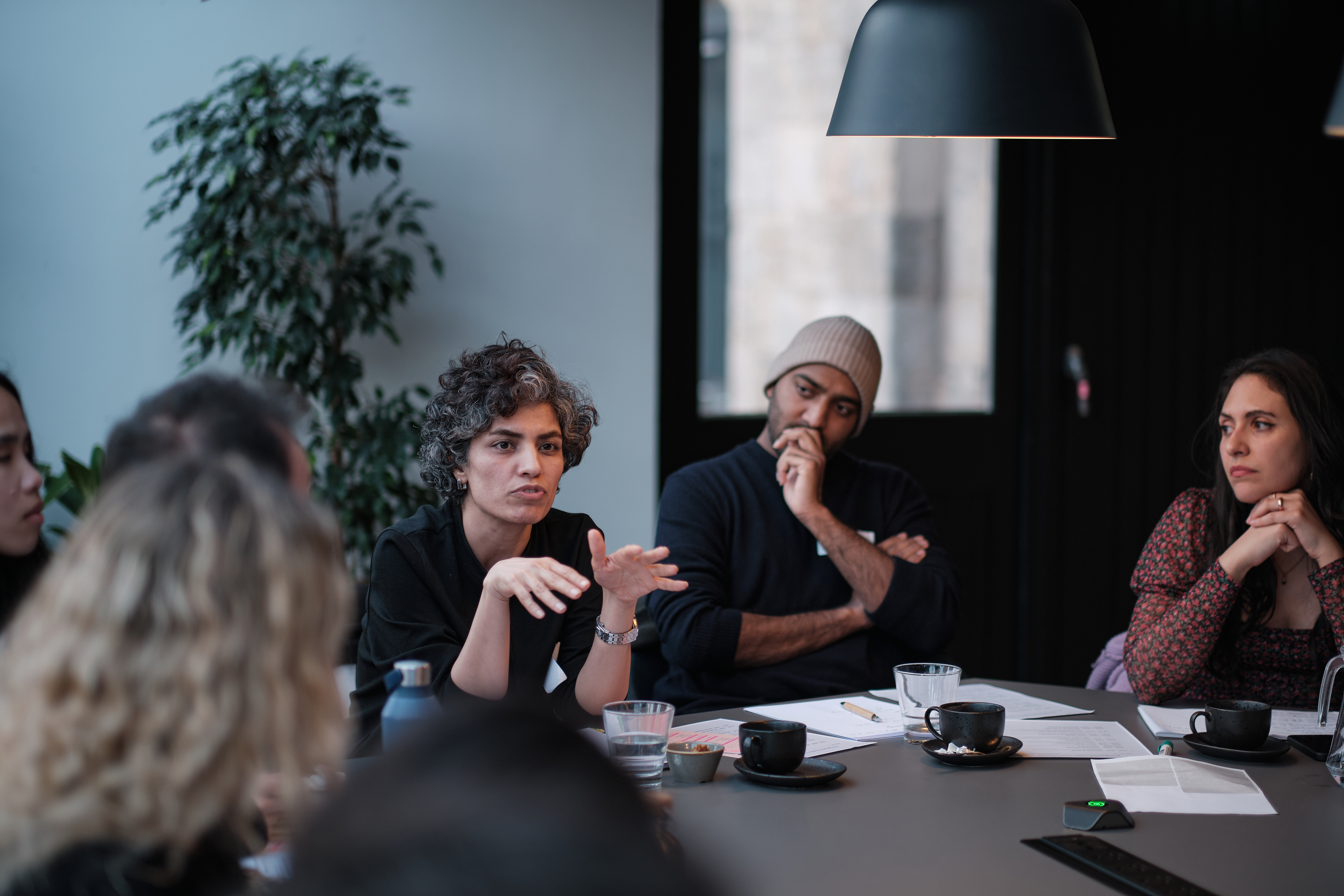 A group of people in discussion around a table, prominent people are a woman with black/grey hair and dark top. Beside her a dark skinned man with white beanie.