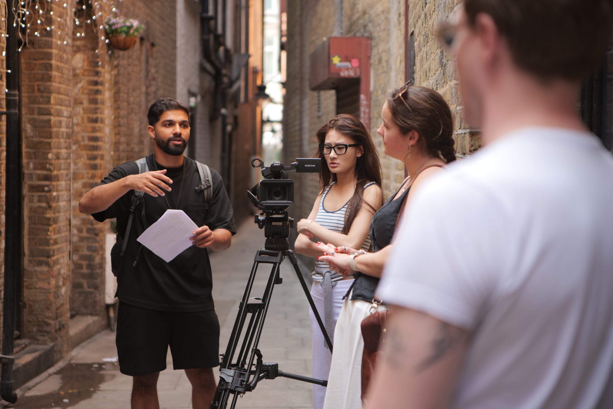 An outdoor shoot in an alley. A director standing next to a camera gives directions to 3 members of his production crew