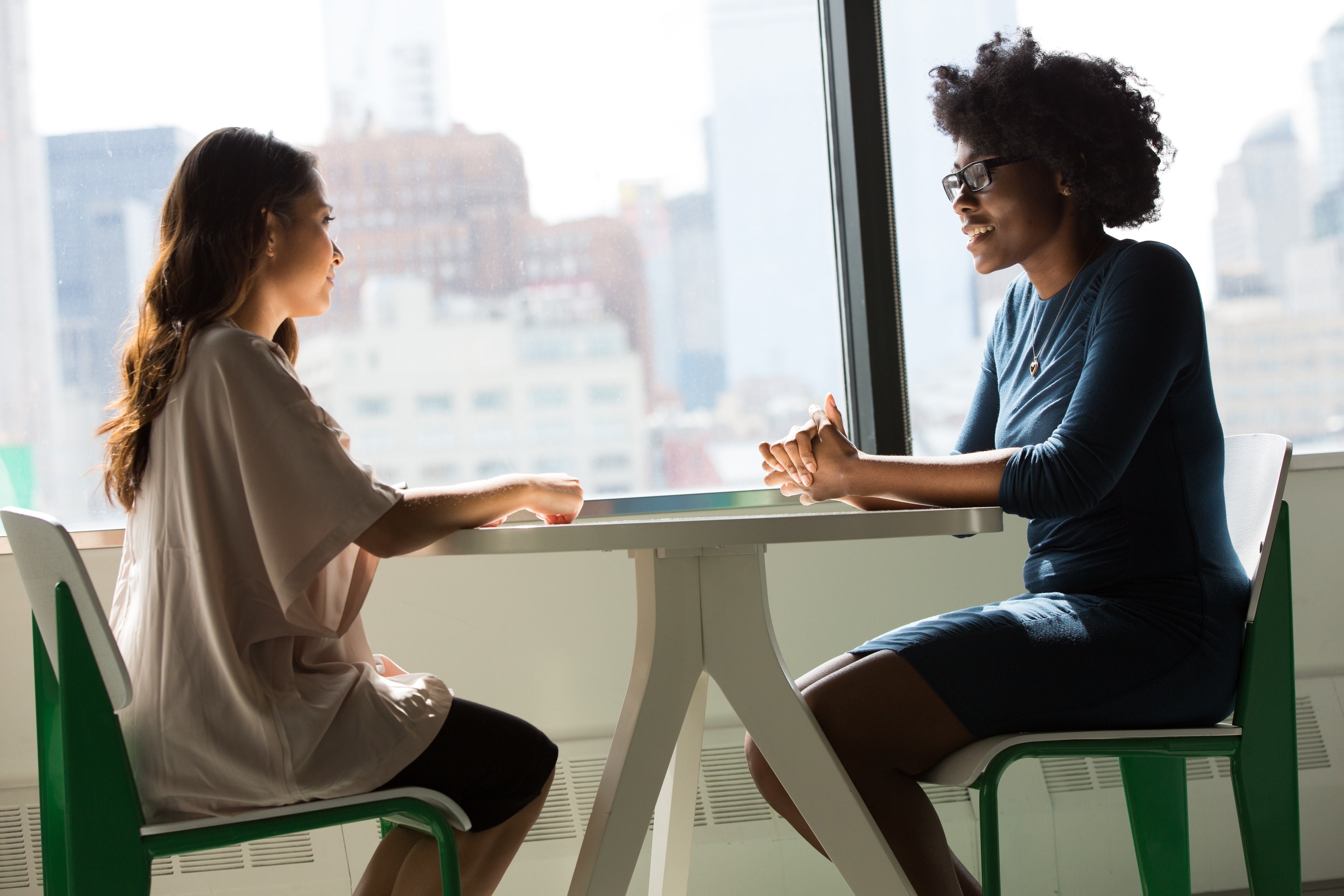 Two women in conversation sat across from one another at a table, in front of a window overlooking a cityscape.
