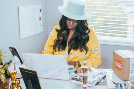 A woman wearing a yellow blouse and white hat sits working at a desk